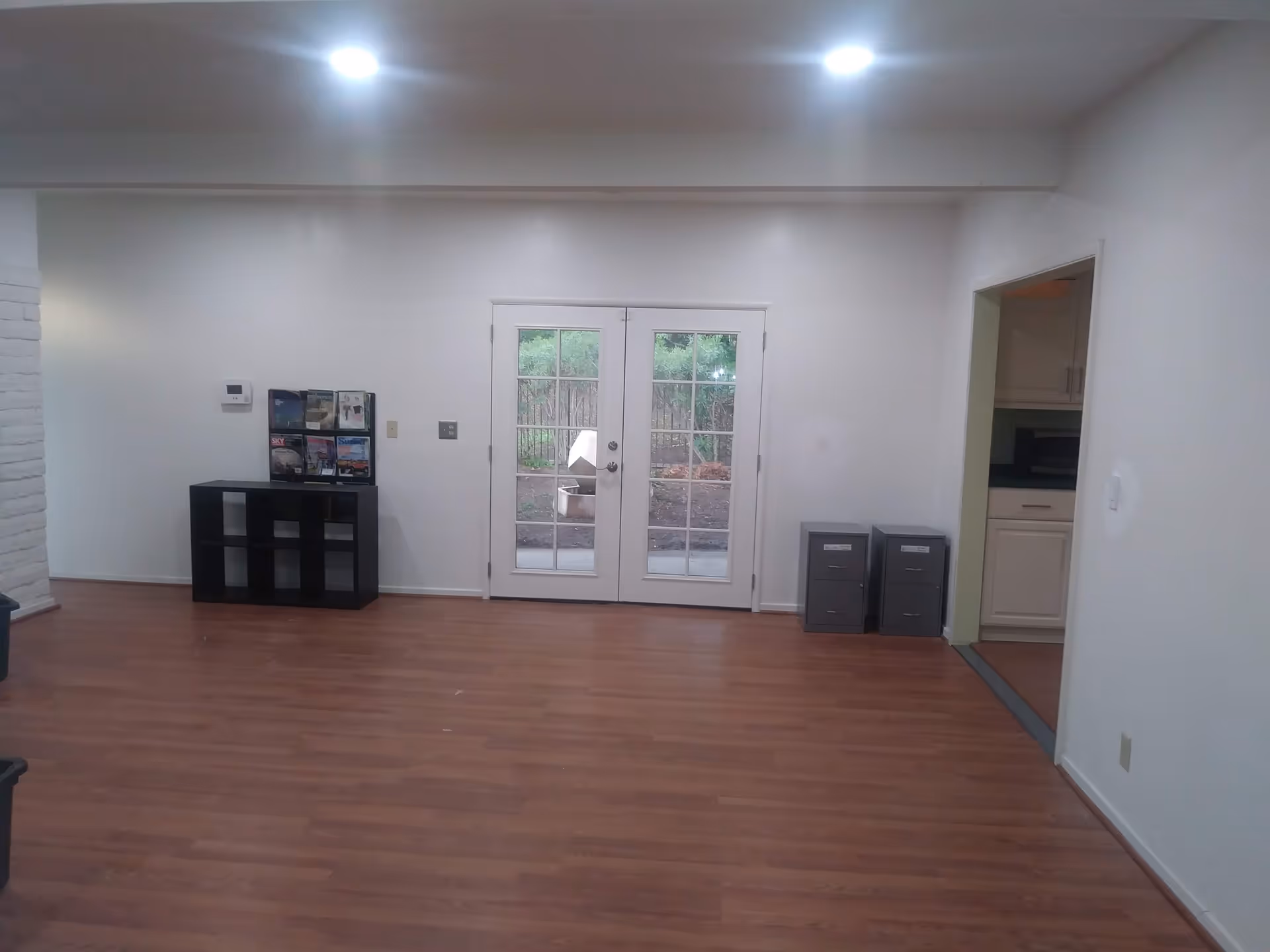 A room with wooden flooring and white walls featuring a set of double glass doors leading outside. There is a small black shelving unit with magazines on top against the left wall and two gray filing cabinets near the doorway to a kitchen area on the right.