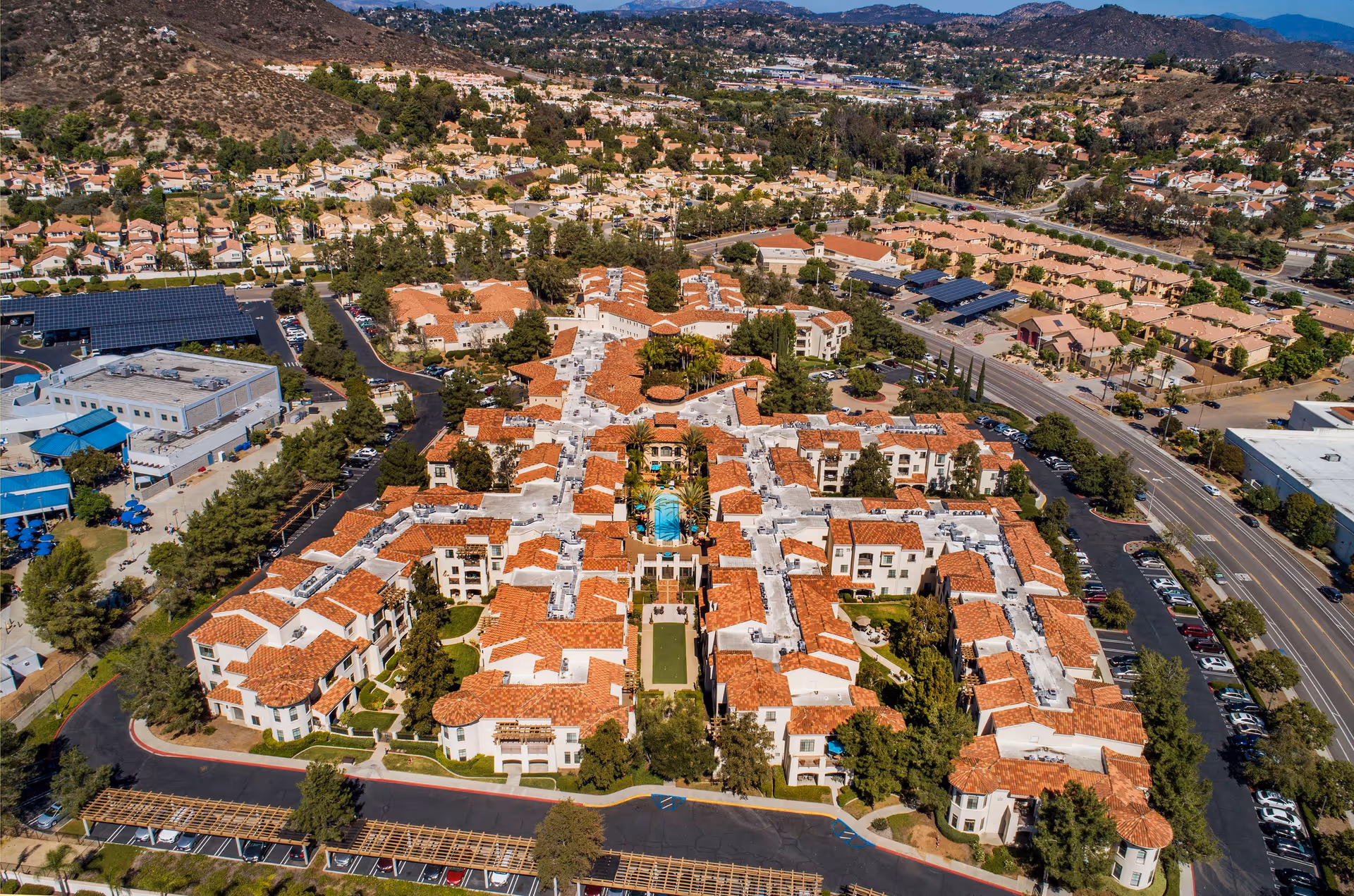 Aerial view of La Vida Real senior living complex with terracotta-roofed buildings clustered around a central pool and courtyard, surrounded by roads and neighborhood hills.