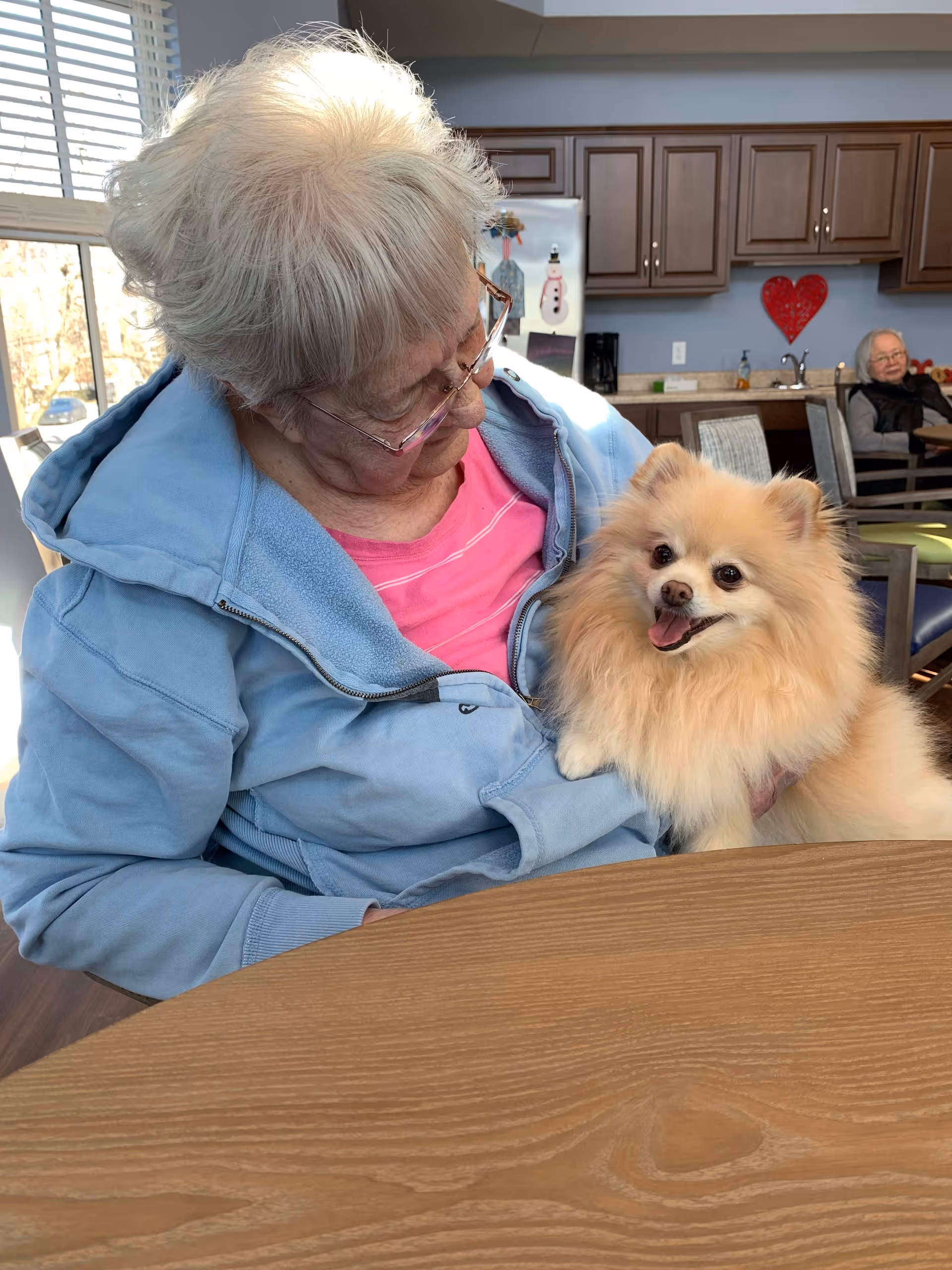 An elderly woman wearing glasses, a pink shirt, and a light blue jacket is sitting at a wooden table holding a small fluffy dog. The dog is looking at the camera with its mouth open, appearing happy. In the background, there is a kitchen area with brown cabinets, a refrigerator decorated with a snowman magnet, and another elderly woman sitting at a table.