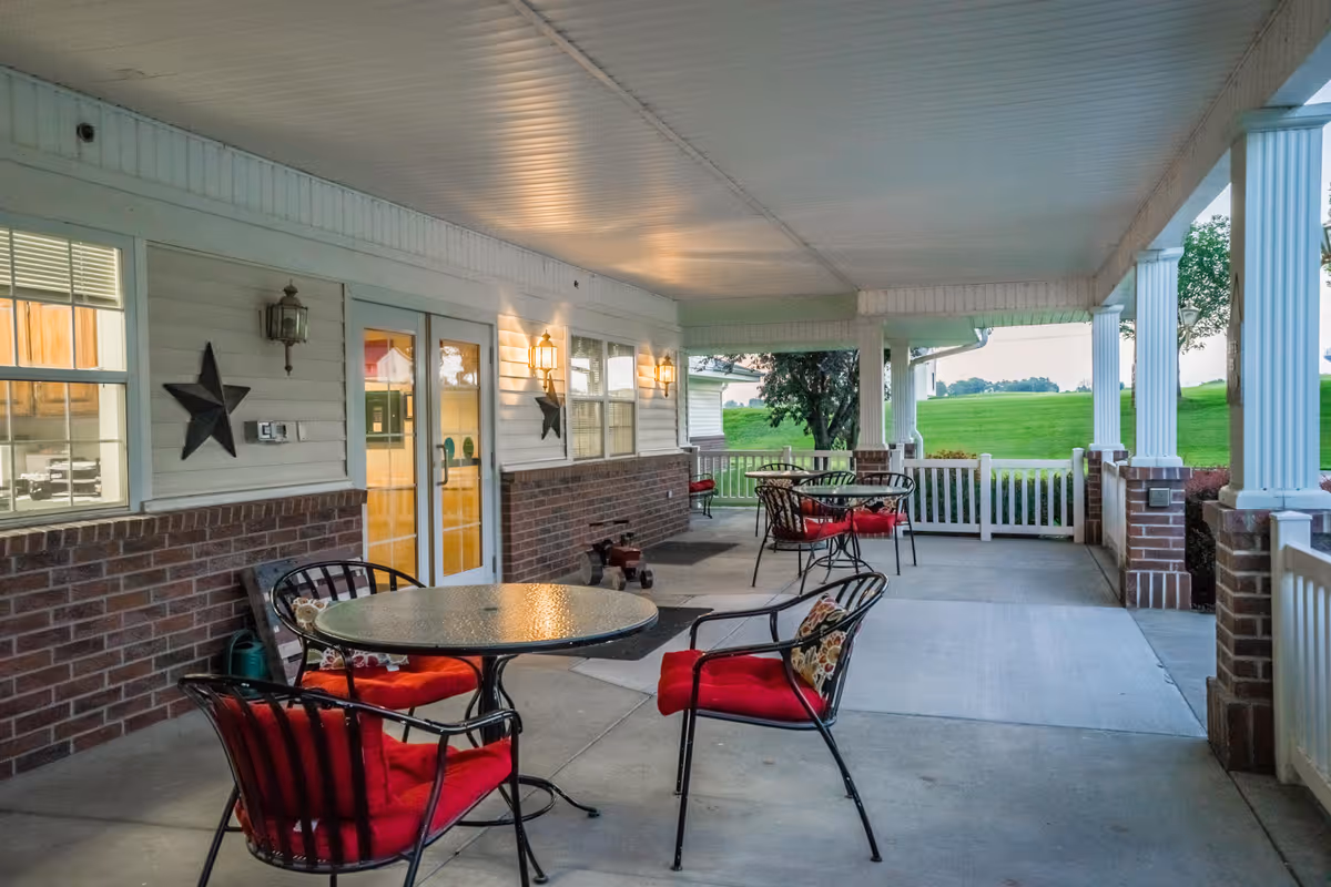 Covered outdoor patio area with round glass tables and black metal chairs with red cushions. The patio has white pillars and railing, brick half-walls, and is adjacent to a building with white siding and windows. Green grass and trees are visible in the background.