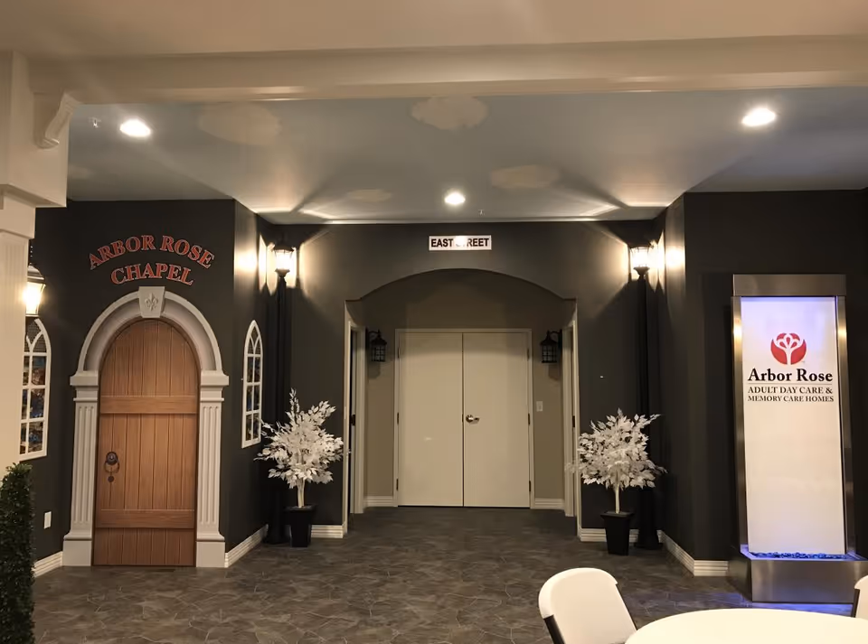 Interior hallway area of Arbor Rose of Tolono featuring a decorative wooden door labeled Arbor Rose Chapel on the left, two white potted plants flanking a double door in the center, and a tall illuminated sign on the right displaying the Arbor Rose logo and text 'Adult Day Care & Memory Care Homes'. The floor has a stone tile pattern and the walls are painted dark gray with white trim.