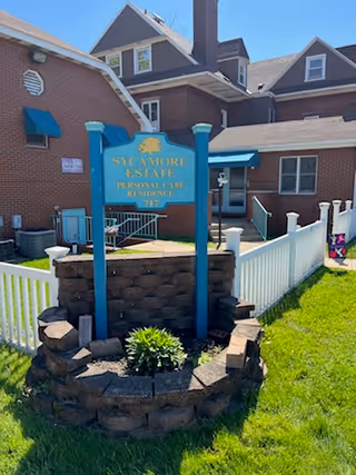 Exterior view of Sycamore Estate Personal Care Residence showing a brick building with multiple windows and a blue sign in front. The sign is mounted on blue posts and surrounded by a small stone planter with greenery. A white picket fence runs along the side of the building, and the grass is green and well-maintained under a clear sky.