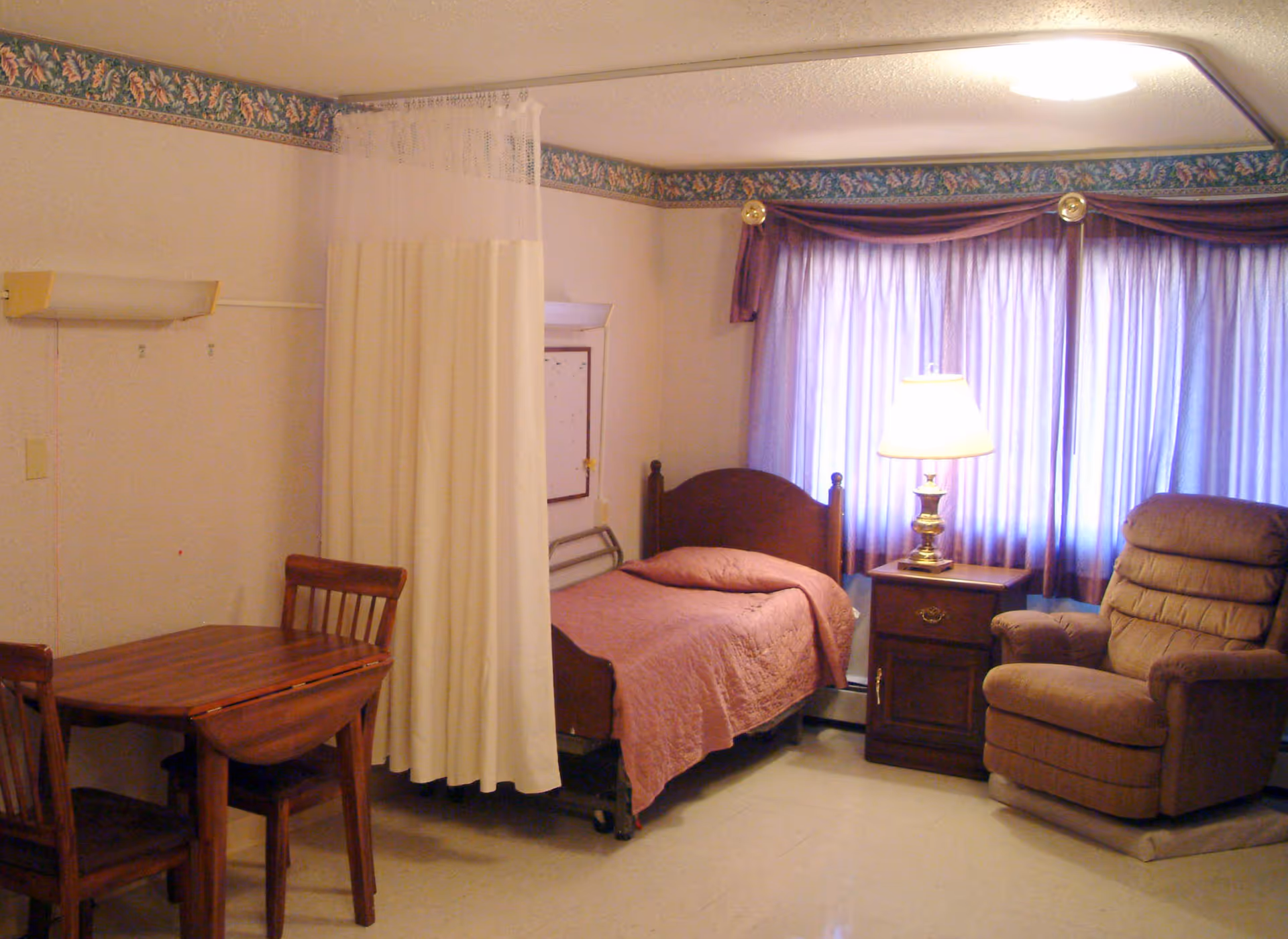A senior care resident room with a single bed covered by a pink bedspread, a nightstand with a lamp, a recliner, a small dining table and a privacy curtain.