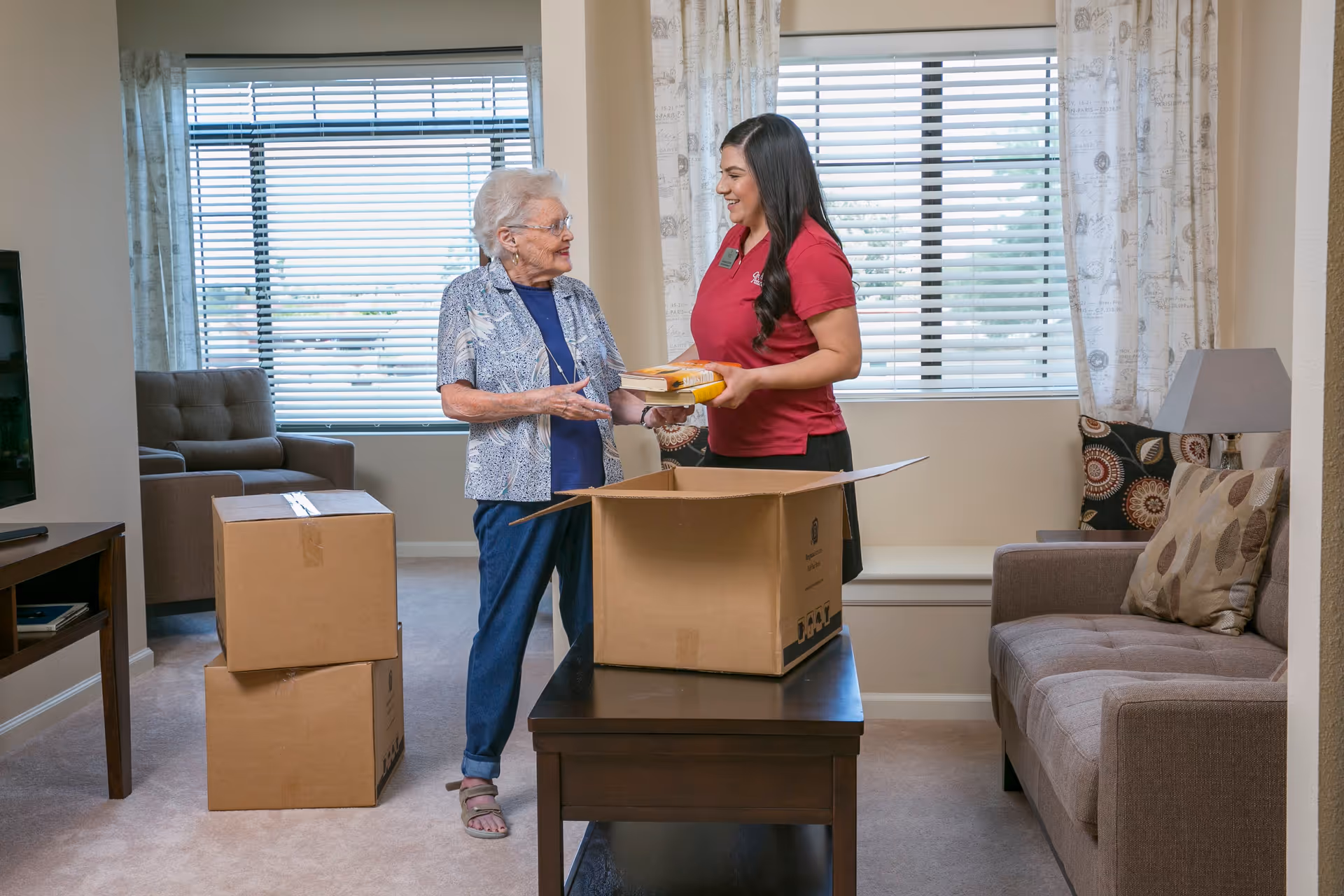 An elderly woman and a younger woman in a red shirt are standing in a living room with beige walls and carpet. The elderly woman is handing a book to the younger woman who is smiling and holding two books. There are two cardboard boxes, one open on a coffee table and two stacked on the floor. The room has a brown sofa with patterned pillows, a lamp, and windows with blinds and patterned curtains.