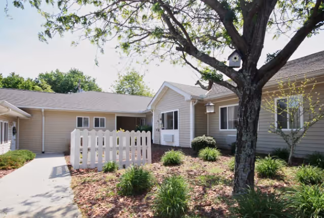 Exterior view of a single-story building with beige siding and a gray roof, surrounded by landscaped garden beds with green shrubs and a large tree with a birdhouse. A white picket fence and a concrete walkway lead to the entrance.
