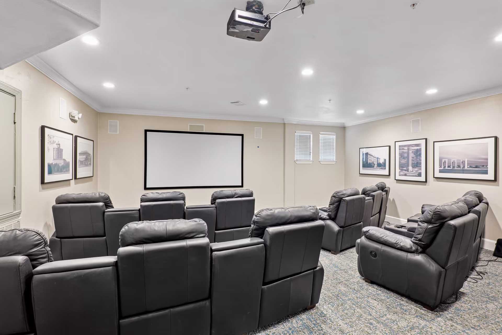 A small theater room with two rows of black leather recliner chairs facing a large white projection screen on a beige wall. The room has recessed ceiling lights, framed pictures on the walls, and a projector mounted on the ceiling.