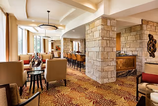 Spacious interior lounge area with stone pillars, patterned carpet, multiple seating arrangements including armchairs and bar stools, large windows allowing natural light, and modern ceiling lights.