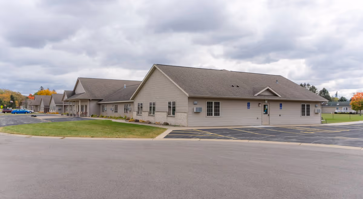 Exterior view of a single-story senior living facility building with beige siding and a gray shingled roof. The building has multiple windows and a small entrance door with handicap signs on either side. There is a parking lot with marked spaces and a grassy area in front. The sky is cloudy.