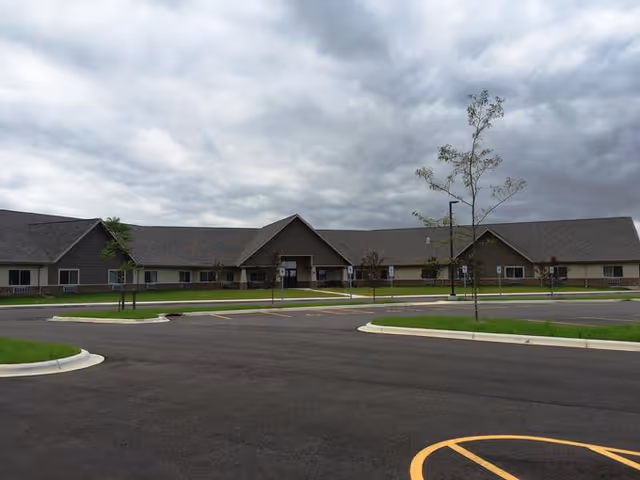 Exterior view of a single-story building with a large parking lot in front, under a cloudy sky. The building has a pitched roof and multiple windows, with a main entrance in the center.