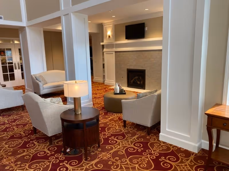 A cozy living room area with beige armchairs and a loveseat arranged around a round ottoman in front of a fireplace with a mounted TV above it. The room features a patterned red carpet, white paneled walls, and a wooden side table with a lamp.