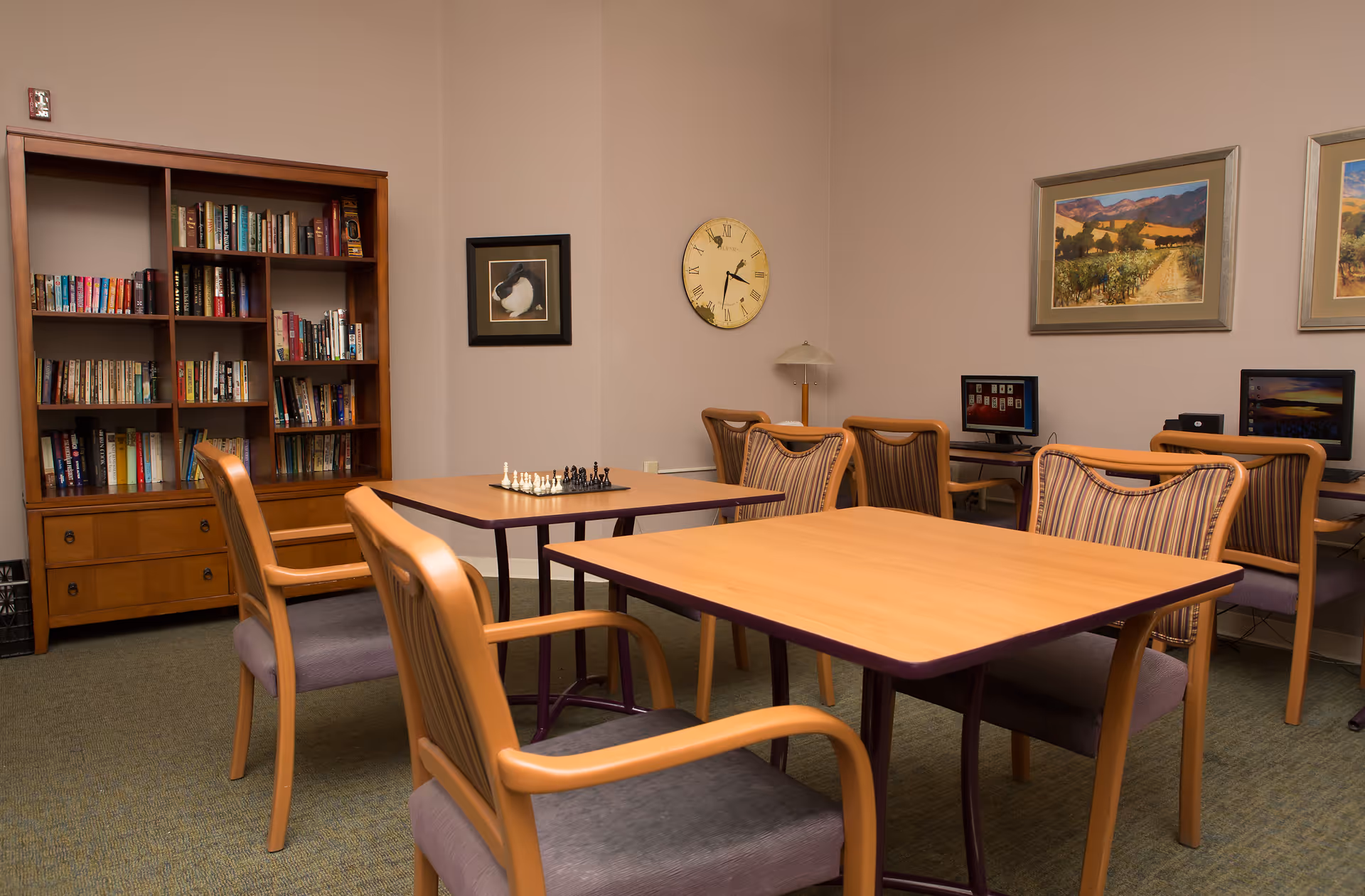 A quiet room with wooden tables and chairs arranged for activities. One table has a chessboard set up. There is a wooden bookshelf filled with books against the wall, a clock, framed pictures, and two computer stations with chairs along the far wall.