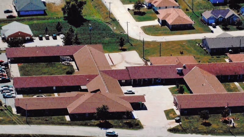 Aerial view of The Village Retirement Community showing multiple single-story buildings with red roofs arranged around paved driveways and green lawns. Several cars are parked near the buildings and the surrounding area includes additional houses and streets.