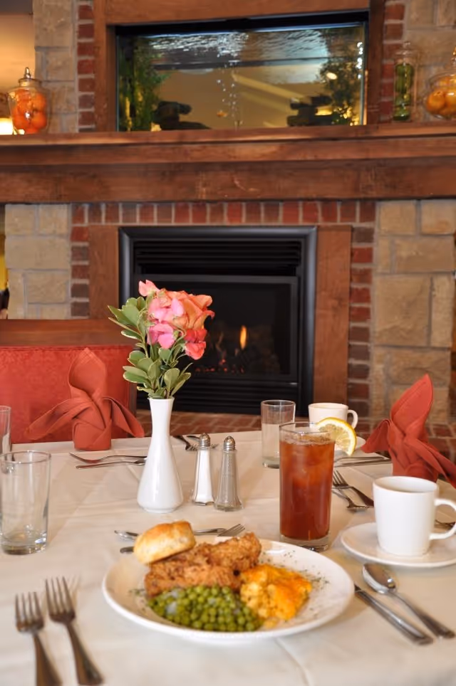 A dining table set with a plate of fried chicken, green peas, a biscuit, and macaroni and cheese. The table also has a glass of iced tea with a lemon slice, a white coffee cup, salt and pepper shakers, a white vase with pink and orange flowers, and folded red napkins. In the background, there is a stone and brick fireplace with a fire burning and a large fish tank above it.