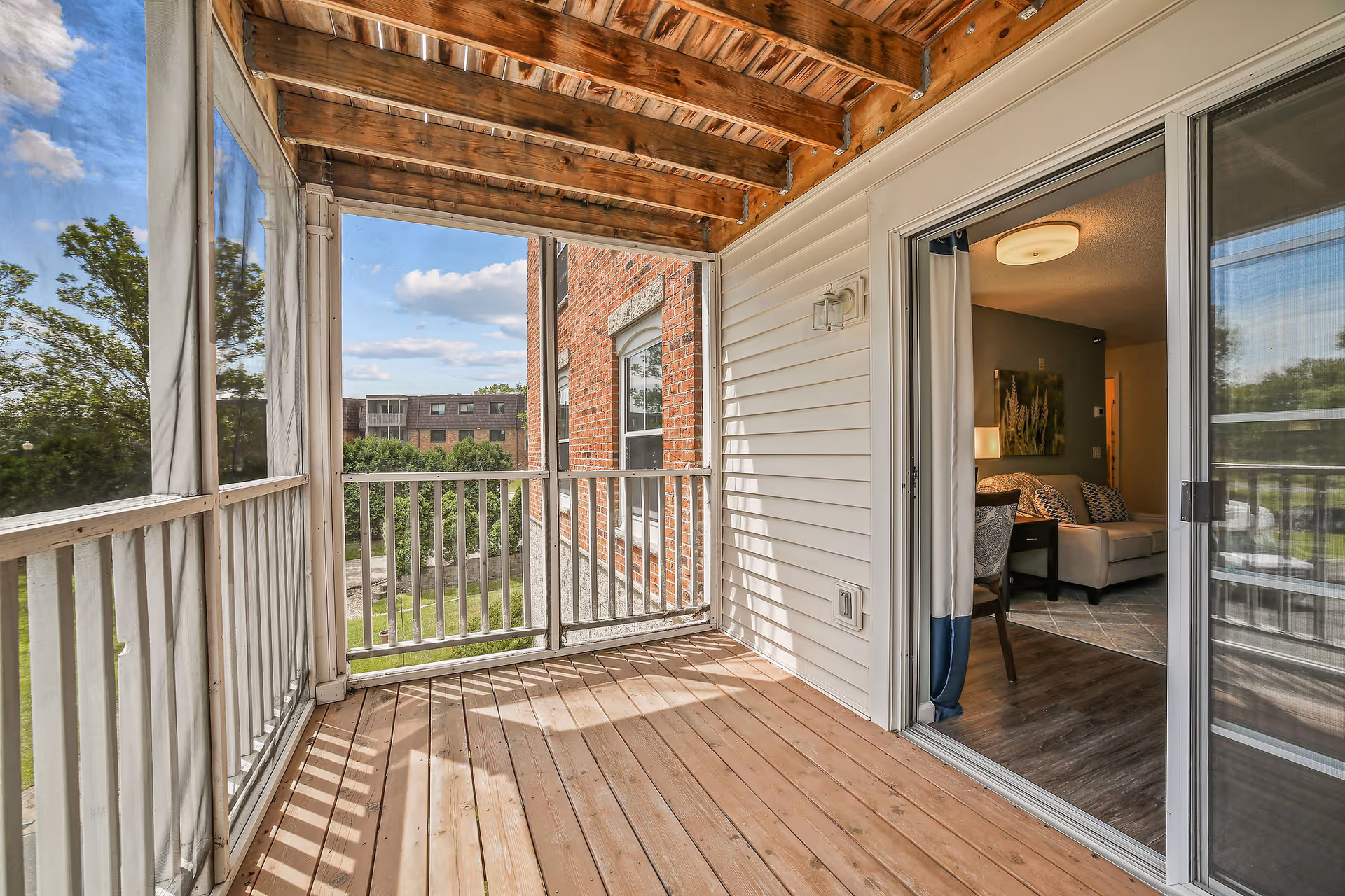 A screened-in wooden balcony with a view of trees and a neighboring brick building. The balcony has a wooden floor and ceiling with exposed beams. A sliding glass door opens into a living room area with a sofa, chair, lamp, and wall art.
