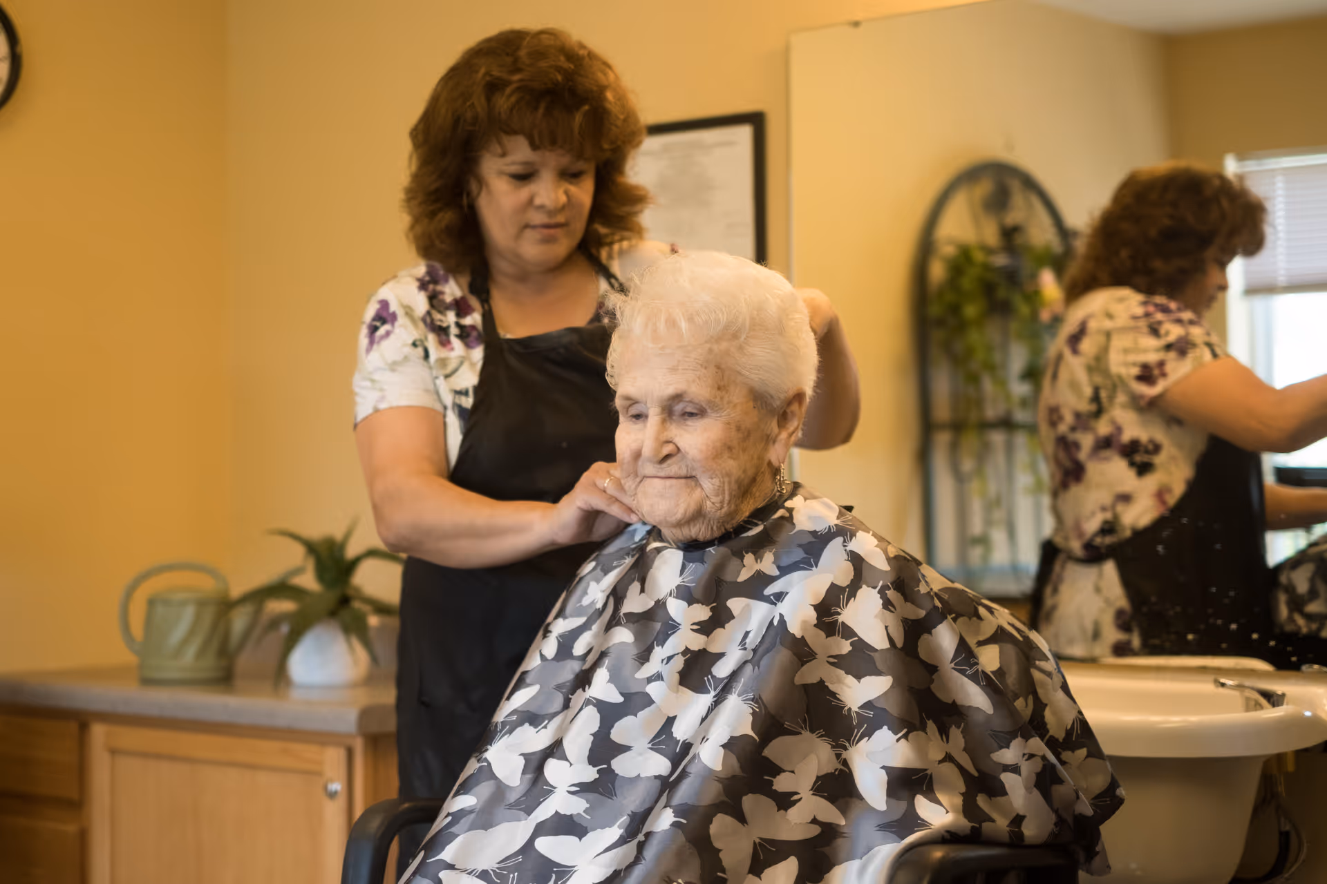 An elderly woman with white hair is sitting in a chair wearing a black and white butterfly-patterned cape while a woman with curly brown hair and a floral shirt adjusts the cape around her neck in a hair salon setting. A mirror reflects the woman adjusting the cape, and there is a sink and some plants in the background.