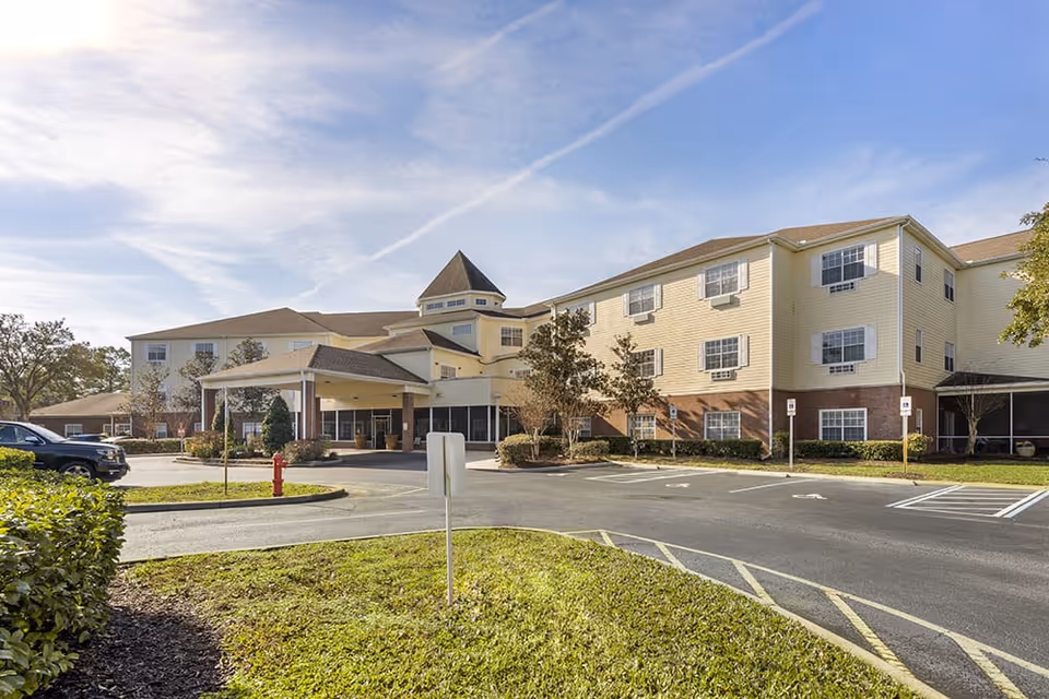 Exterior view of Brookdale Lake Mary senior living facility showing a three-story building with beige siding and brick accents, a covered entrance, parking spaces, and landscaped greenery under a partly cloudy sky.