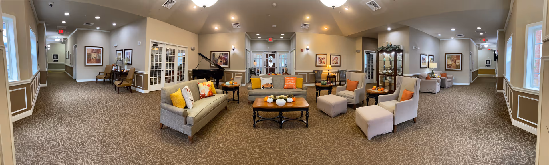 A spacious and well-lit common area in a senior living facility featuring comfortable seating including sofas and armchairs with colorful cushions, a coffee table with decorative pumpkins, framed artwork on the walls, a piano in the corner, and large windows letting in natural light.