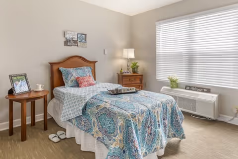 A neatly made single bed with a patterned blue and white bedspread and multiple pillows in a bright room. Next to the bed is a wooden nightstand with a lamp, a small plant, and a book. There is a round wooden side table with a framed photo and a cup. A large window with white blinds lets in natural light, and a white heater is positioned below the window. The walls are light-colored, and the floor is carpeted.
