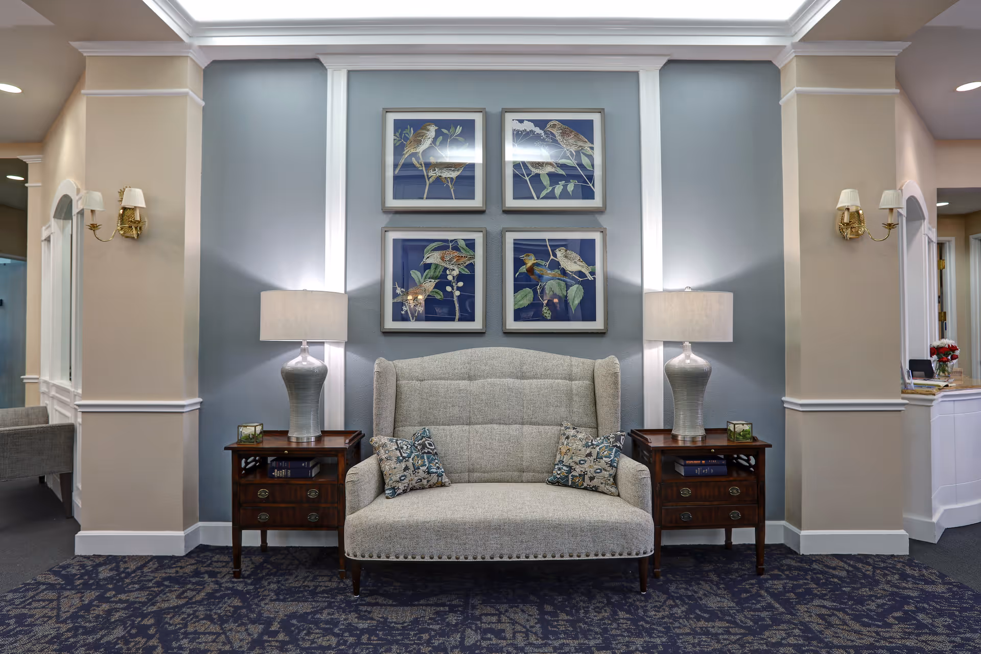 A cozy seating area in a healthcare facility with a light gray upholstered loveseat flanked by two wooden side tables, each holding a silver lamp with a white shade. Above the loveseat are four framed pictures of birds on a blue background. The walls are painted in soft beige and blue tones, and the floor is covered with a patterned blue carpet. Wall sconces with white shades are mounted on the beige walls on either side.