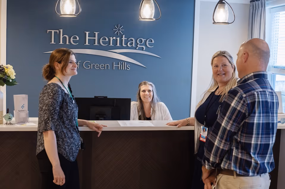 Reception area at The Heritage of Green Hills with a receptionist sitting behind the desk smiling and three people standing in front of the desk engaged in conversation. The background features a blue wall with the facility's name displayed.