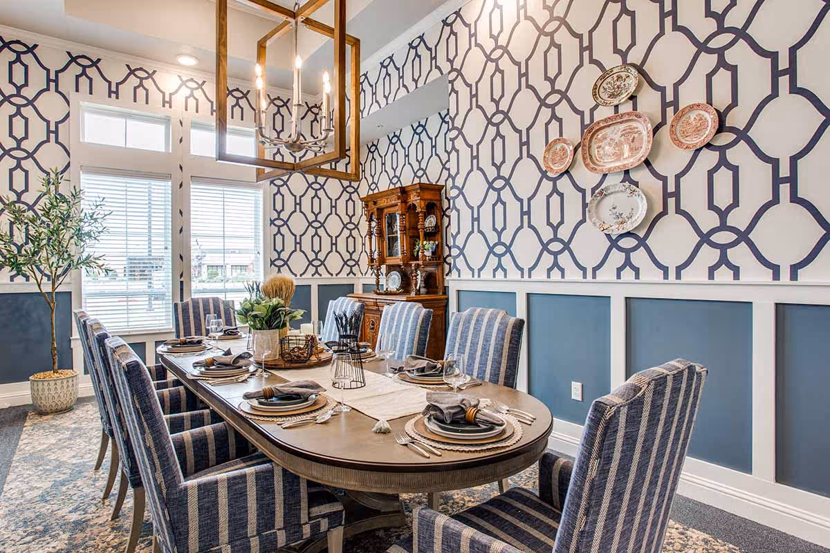 Bright dining room with a long set table, striped upholstered chairs, geometric wallpaper and decorative plates on the wall.