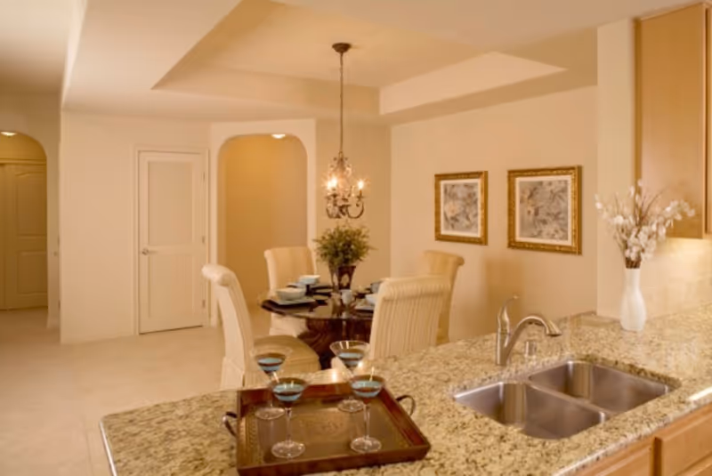Open kitchen with a granite countertop and double sink in the foreground and a dining table with chairs and chandelier in the background.