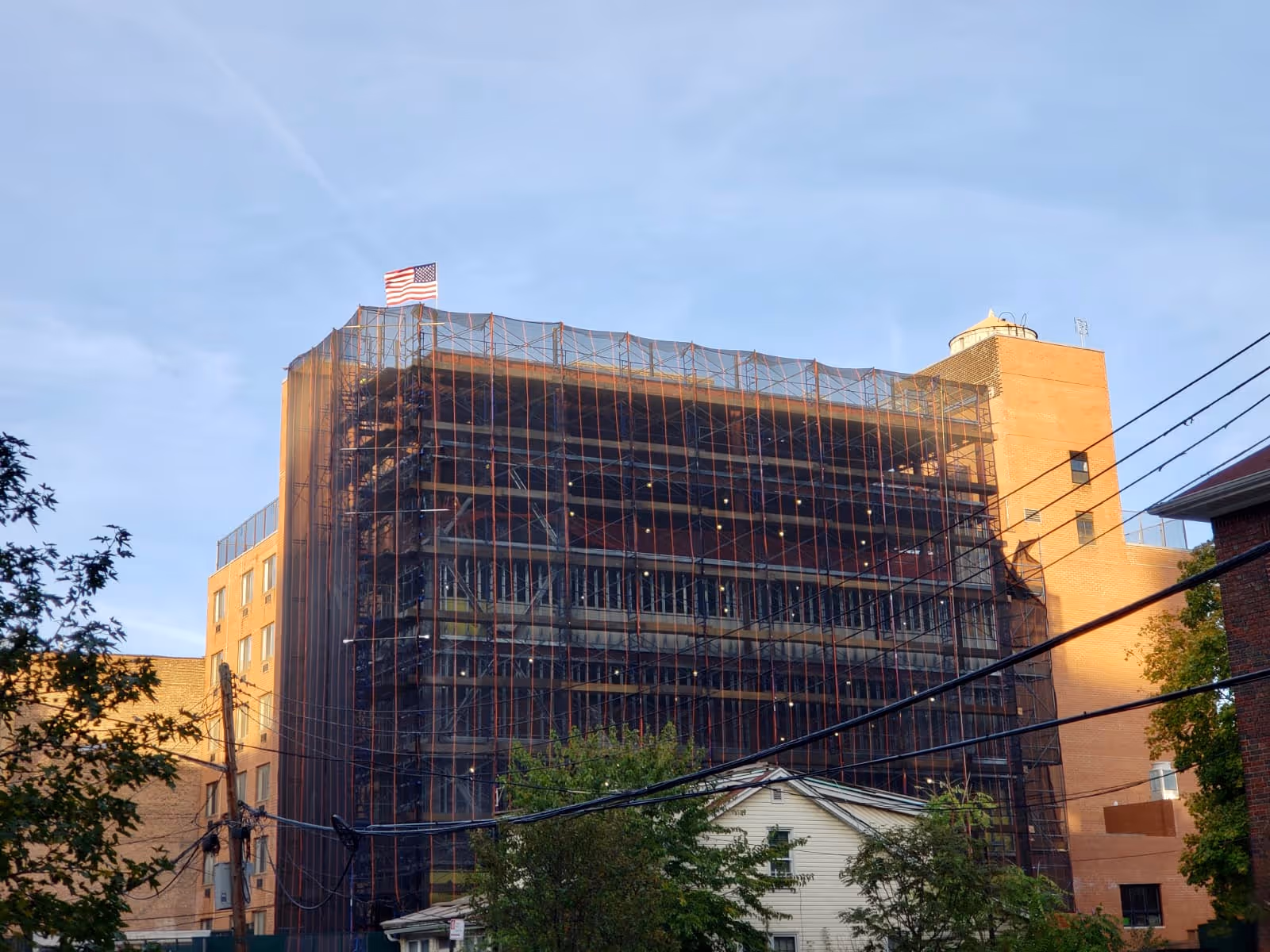 A multi-story brick building under construction or renovation, covered in scaffolding and protective netting. An American flag is flying at the top of the building. Trees and power lines are visible in the foreground under a clear sky.