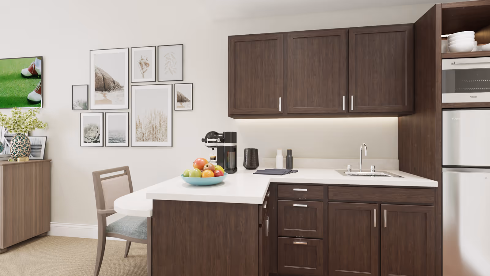 Modern kitchenette with dark wood cabinets, a white countertop island holding a bowl of fruit and a coffee maker, and a chair with framed wall art on the adjacent wall.