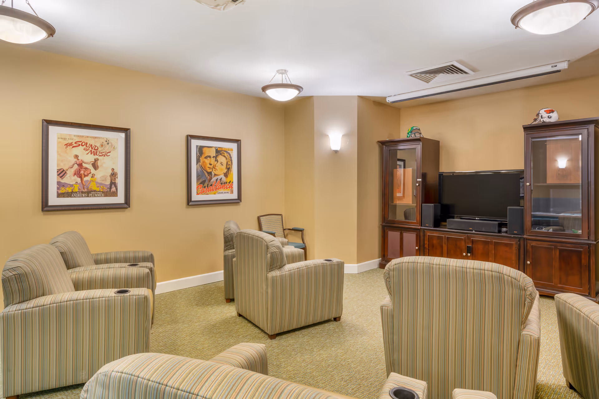 A cozy living room area with several striped armchairs arranged facing a wooden entertainment center with a flat-screen TV and speakers. The walls are painted beige and decorated with framed vintage movie posters of 'The Sound of Music' and 'Casablanca'. The room is softly lit by ceiling lights and a wall sconce.