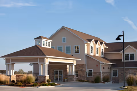 Exterior view of a two-story senior living facility building with beige siding, stone accents, and a covered entrance. The building has multiple windows and a small tower-like structure on the roof. There is a street lamp and landscaped bushes around the paved driveway.