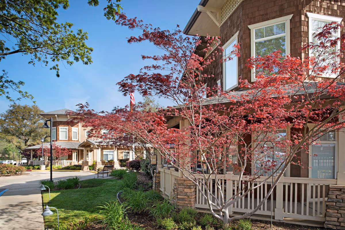 Exterior view of a senior living facility with brown siding and stone accents, surrounded by well-maintained landscaping including green grass, shrubs, and trees with red leaves under a clear blue sky.