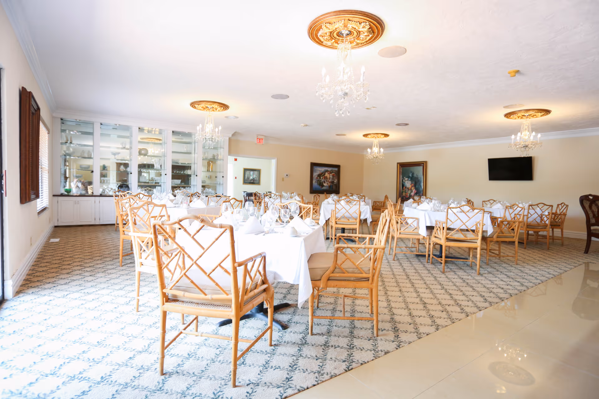 A bright and spacious dining room with multiple tables covered in white tablecloths, set with glassware and napkins. The room features wooden chairs with a lattice design, patterned carpet, chandeliers hanging from the ceiling, framed paintings on the walls, and a flat-screen TV mounted on one wall. Large windows with wooden shutters allow natural light to fill the space.
