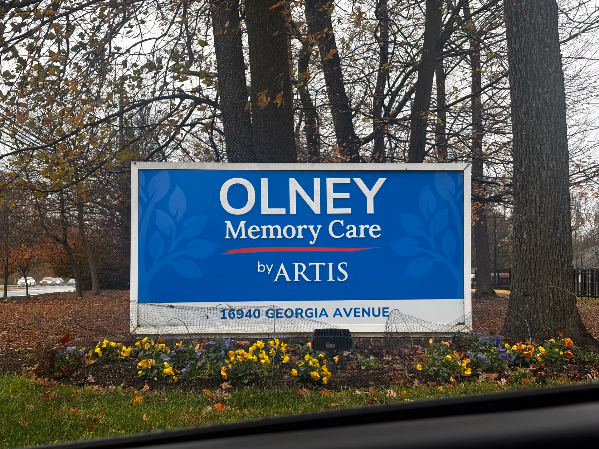 A blue sign reading "OLNEY Memory Care by ARTIS" stands among flowers and trees at 16940 Georgia Avenue.