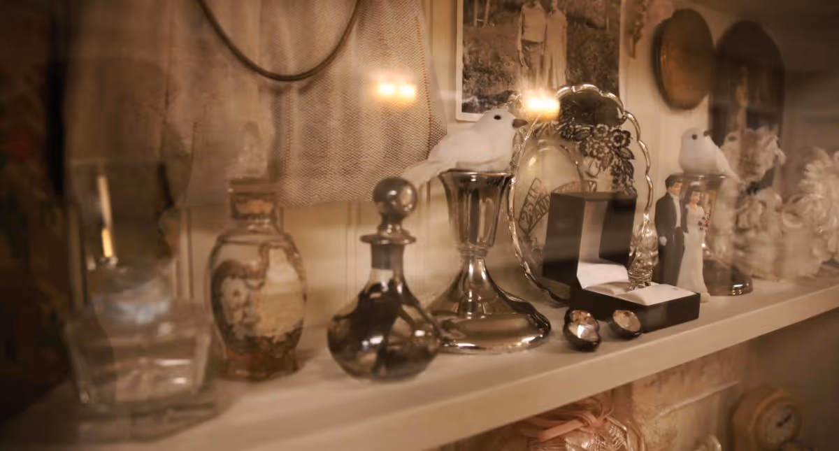 A close-up view of a decorative shelf displaying various vintage items including glass bottles, silver candlesticks with white bird figurines, a small open jewelry box with a ring, and a figurine of a bride and groom, with a sepia-toned photograph and other decorative elements in the background.