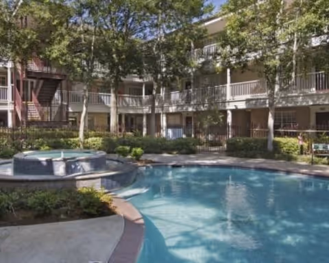 Outdoor swimming pool area surrounded by a multi-story residential building with balconies and trees providing shade.