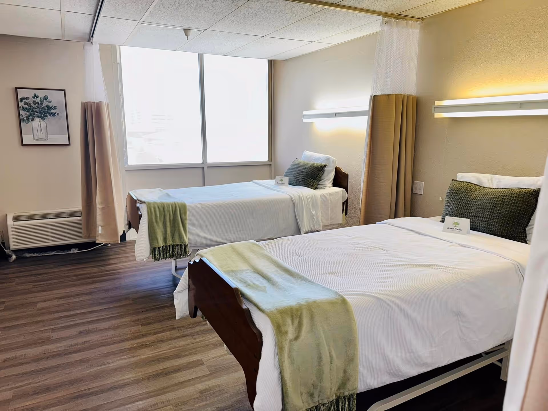 Two neatly made twin beds with green throws and pillows in a bright senior living room with a large window and wood-look flooring.