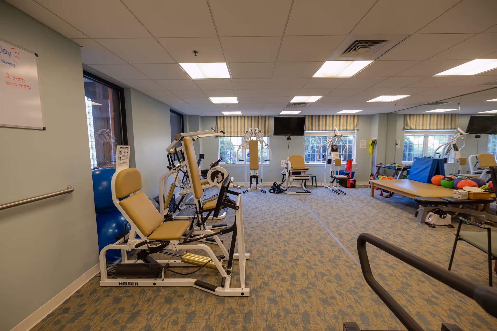 A well-lit exercise room in a senior living facility with various workout machines, exercise balls, and a padded table. The room has large windows with striped curtains, a wall-mounted TV, and a whiteboard on the left wall with some writing. The floor is carpeted and there is a large mirror on the right side.