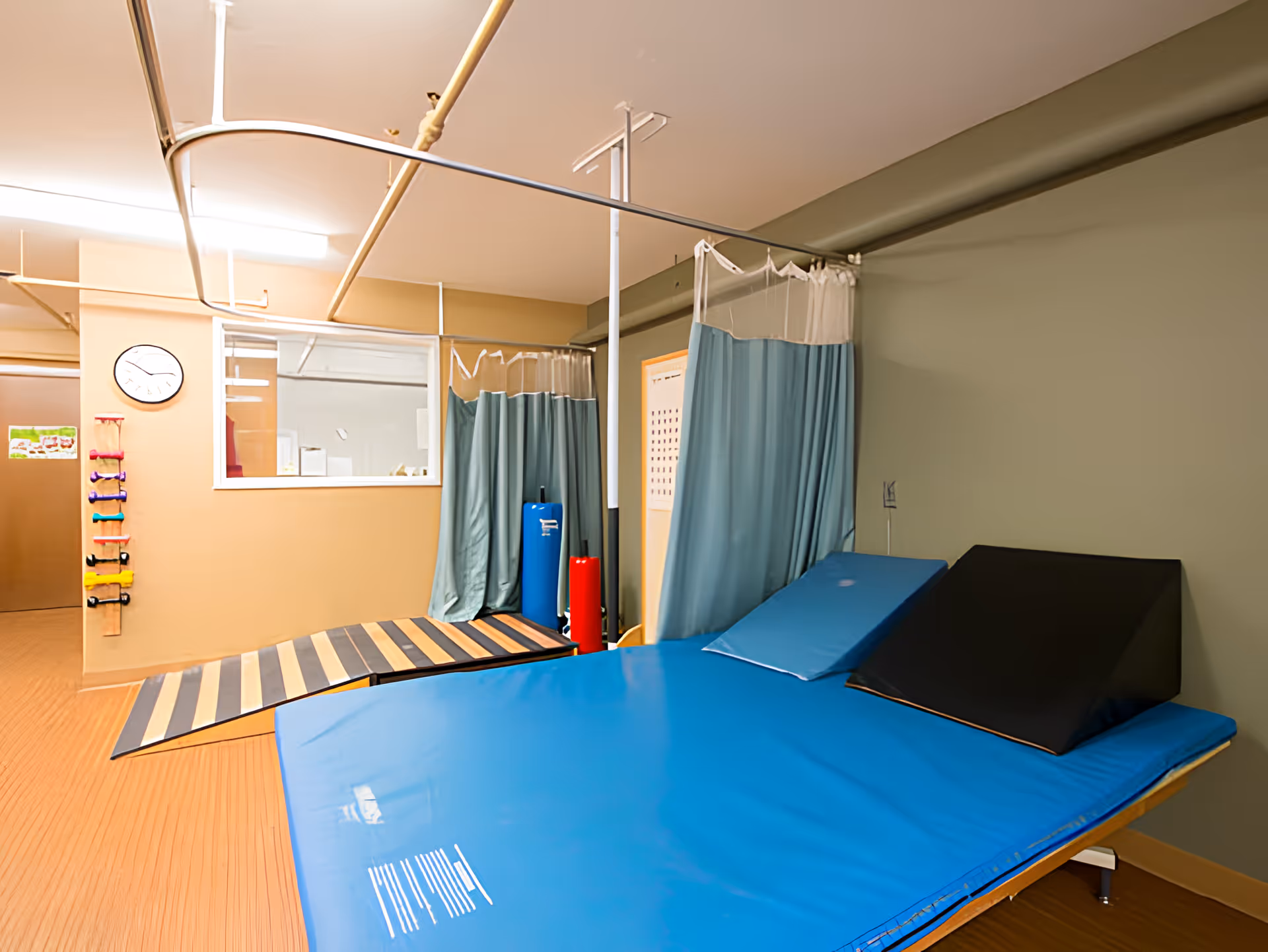 A therapy or rehabilitation room with a large blue padded therapy table, a black wedge cushion, a wooden ramp, and a rack of colorful hand weights mounted on the wall. There are privacy curtains hanging from ceiling tracks and a clock on the wall.