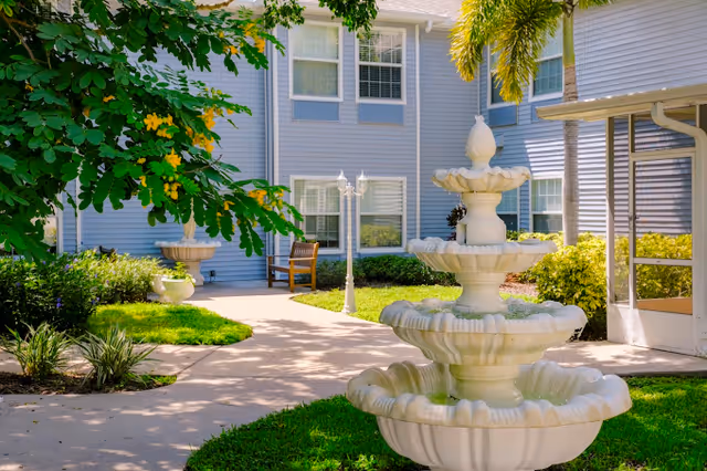 Outdoor courtyard area of a senior living facility with a three-tiered white fountain in the foreground, surrounded by green grass and plants. The background shows a light blue building with white-trimmed windows, a wooden bench, and a white lamp post under a sunny sky.
