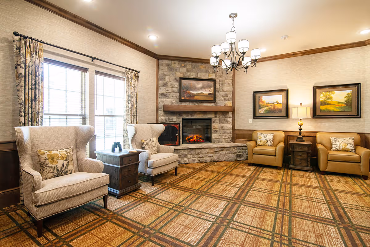 A cozy living room with a stone fireplace in the center, flanked by two beige wingback chairs with floral cushions on the left and two tan armchairs with floral cushions on the right. Between the chairs are wooden side tables, one with a lamp. The room has large windows with patterned curtains, a chandelier hanging from the ceiling, and framed landscape paintings on the walls. The floor is covered with a patterned carpet.