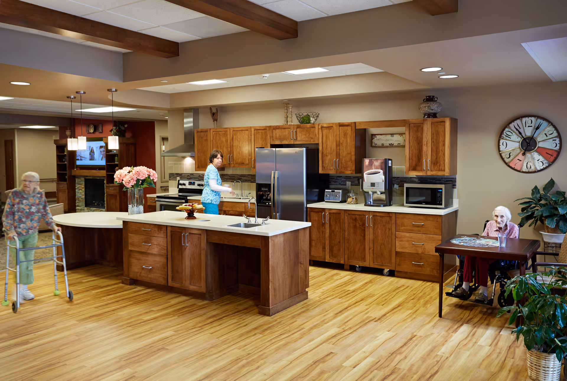 A spacious kitchen area in a senior living facility with wooden cabinets, a large island with a sink, and modern appliances including a refrigerator, microwave, and toaster. Two elderly women are present; one is using a walker and walking near the island, while the other is seated at a small table with a puzzle. A woman in blue scrubs is standing near the sink. The room has wooden flooring, ceiling beams, and a large colorful clock on the wall.