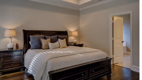 A cozy bedroom with a large dark wooden bed frame and headboard, neatly made with a beige quilt and multiple decorative pillows. There are two matching dark wooden nightstands on either side of the bed, each with a white lamp. The room has light-colored walls and hardwood flooring. An open door leads to an adjacent room with tiled flooring.
