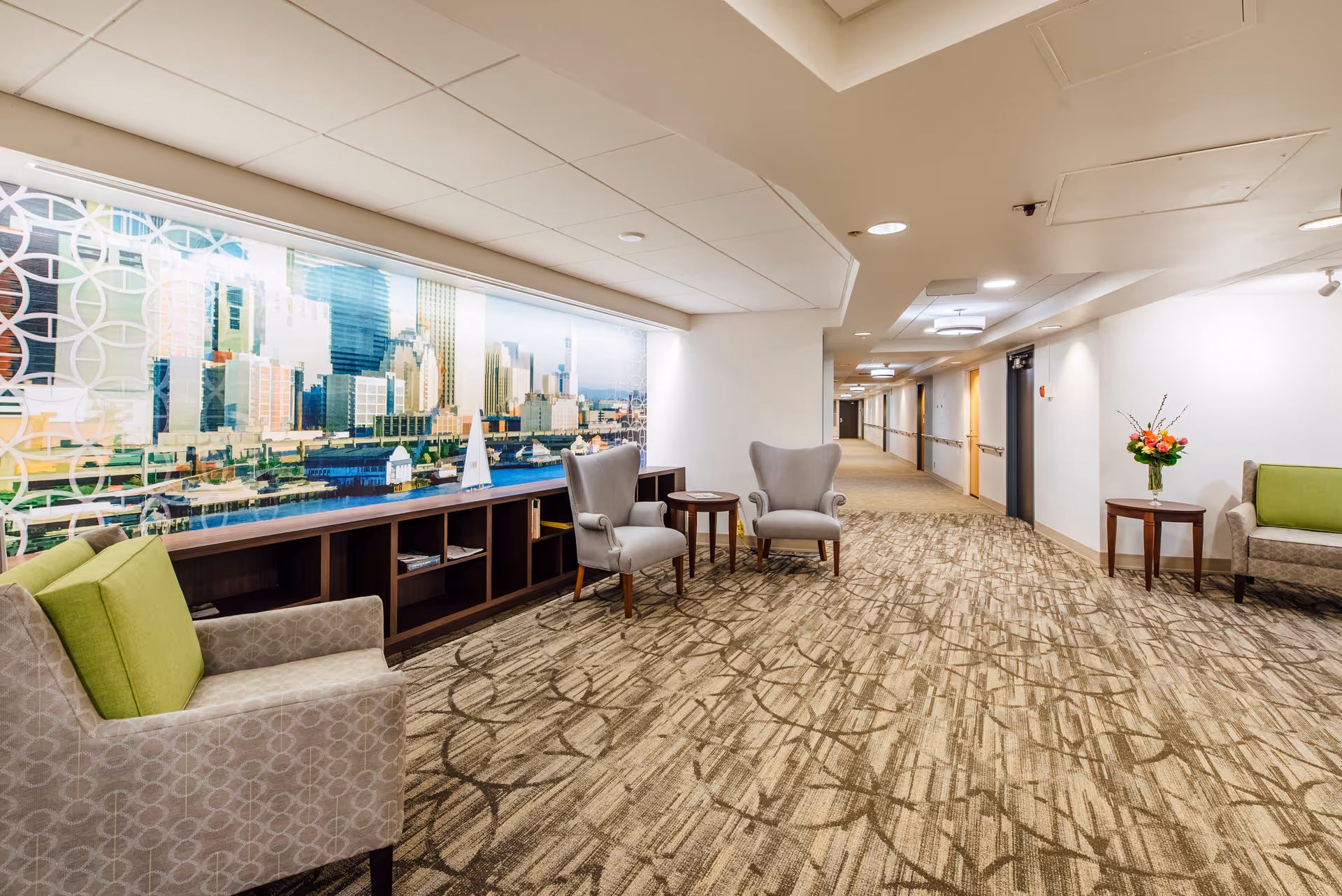 A bright hallway in a retirement community featuring patterned carpet, white walls, and a ceiling with recessed lighting. The hallway includes several comfortable armchairs with green cushions, small wooden side tables, and a large wall mural depicting a cityscape. A vase with flowers is placed on one of the tables near the wall.