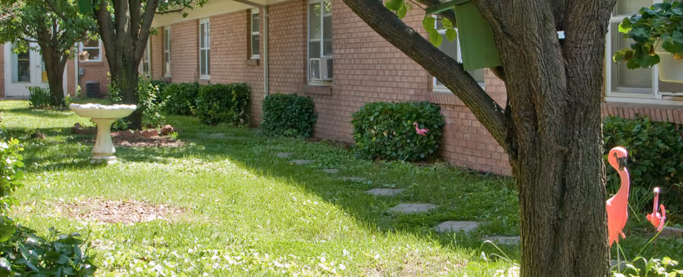 Outdoor garden area with green grass, a tree with a birdhouse, pink flamingo lawn ornaments, a white birdbath, and a brick building with windows in the background.