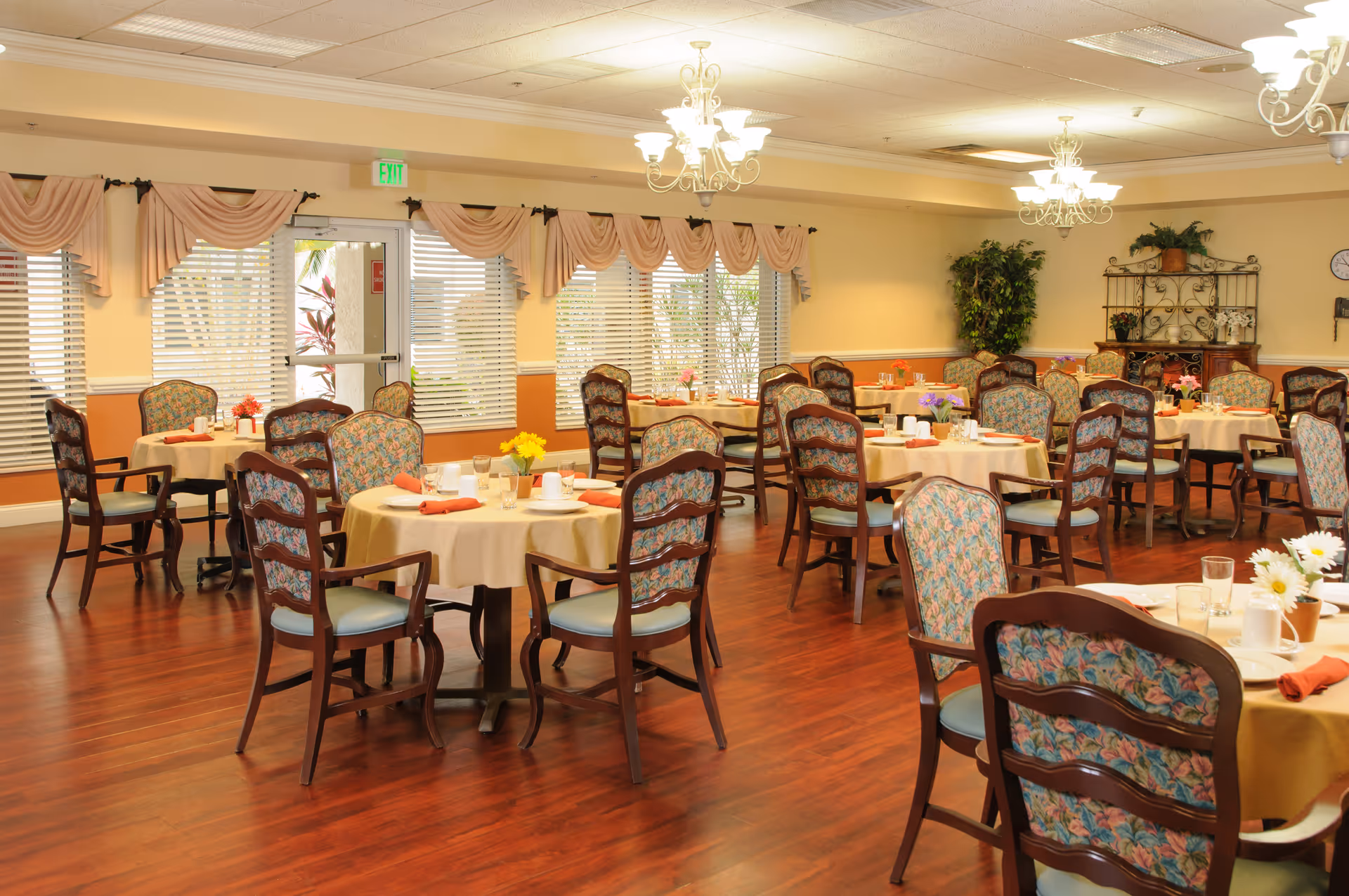 A spacious dining room with multiple round tables covered with beige tablecloths, each set with cups, glasses, and napkins. The chairs have wooden frames with floral patterned upholstery. The room has large windows with blinds and peach-colored valances, wooden flooring, chandeliers hanging from the ceiling, and some plants and decorative shelving along the walls.