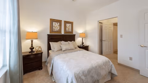 Bright bedroom with a double bed, matching dark nightstands and lamps, framed artwork above the headboard, and an open doorway to an adjacent room.