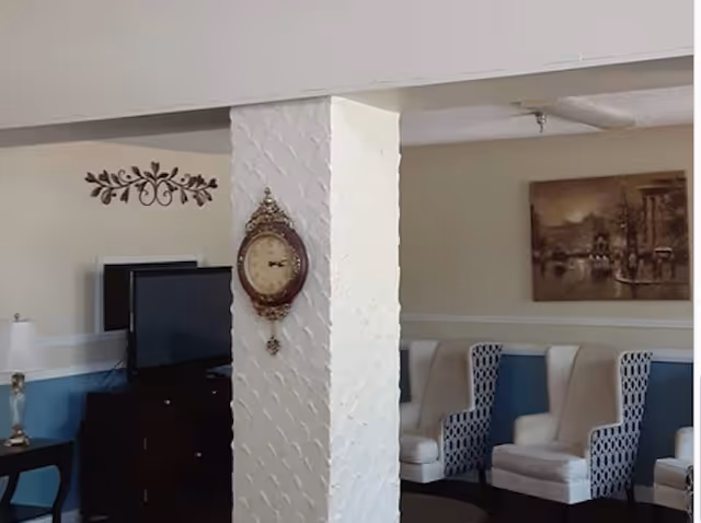 Interior view of a living room area with a white textured pillar in the center featuring a decorative wall clock. To the left, there is a TV on a dark wooden stand and a table lamp on a side table. To the right, there are several upholstered armchairs with patterned backs and a framed painting on the wall.