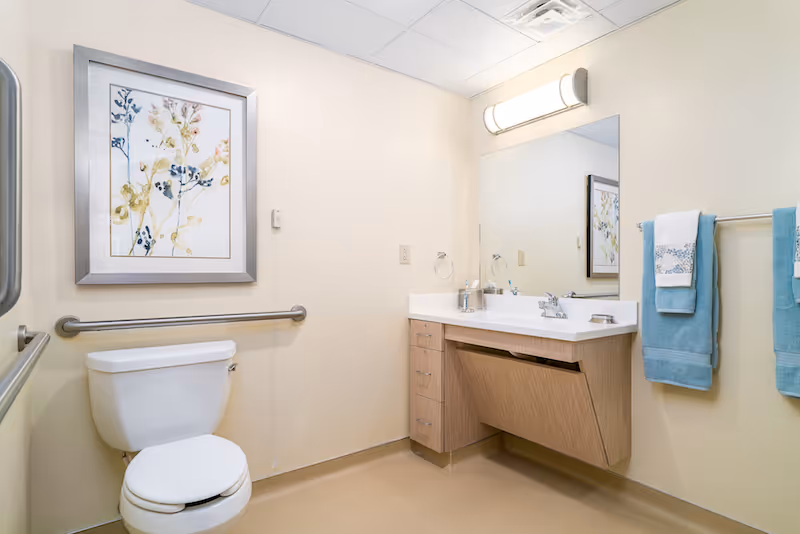 A clean, accessible bathroom with a white toilet, beige walls, and a wooden vanity with a white countertop and sink. There is a large mirror above the sink with a light fixture, two blue towels hanging on a towel rack, and a framed floral artwork on the wall.