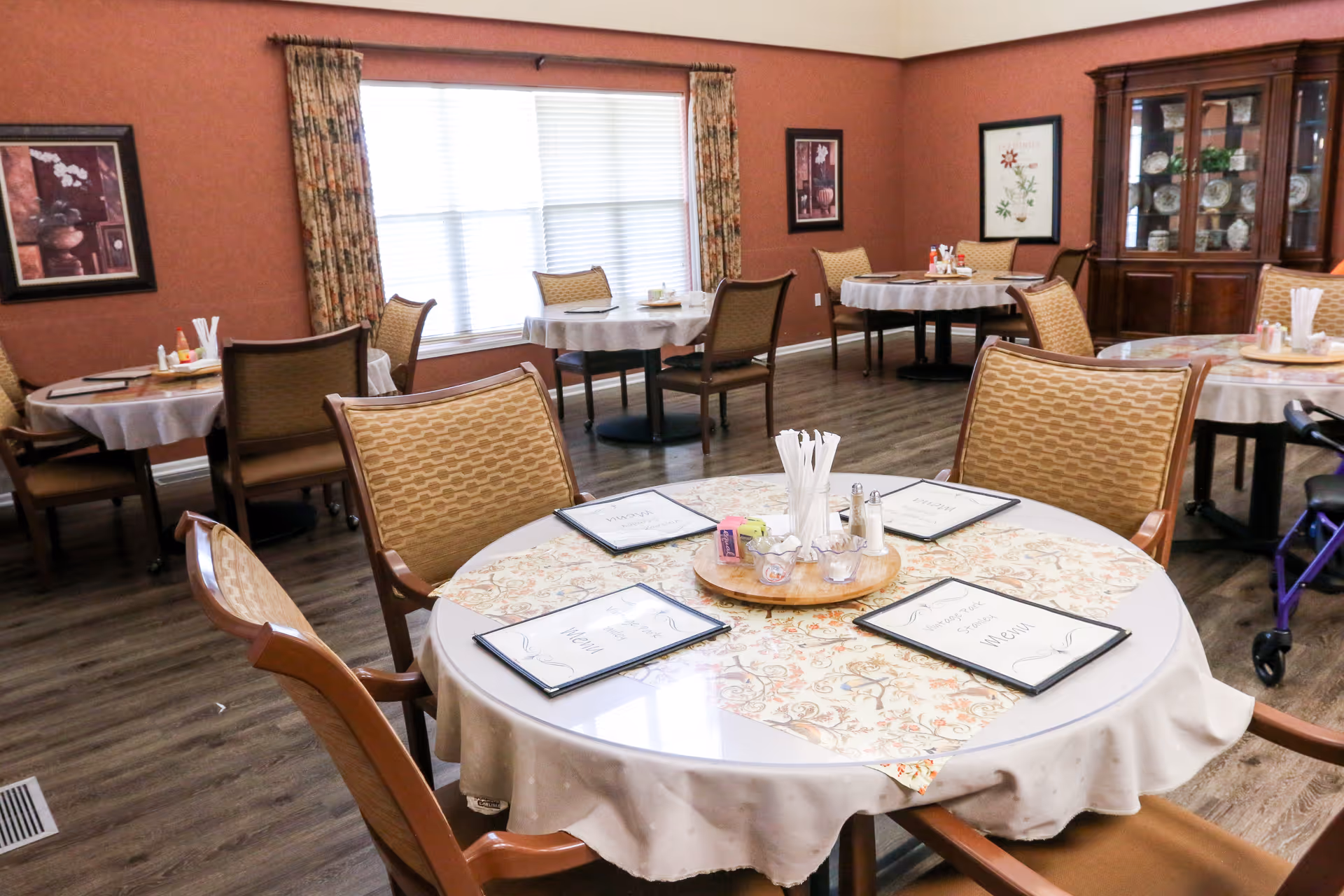 A dining room with round tables covered with tablecloths and menus placed on them. Each table is surrounded by wooden chairs with cushioned seats and backs. The room has wooden flooring, floral curtains on the windows, framed artwork on the walls, and a wooden cabinet displaying dishes and decorative items.
