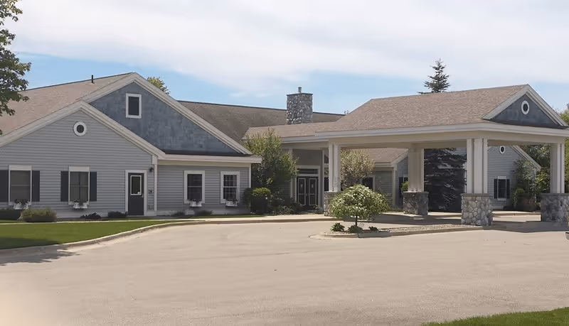 Exterior view of Newberry Assisted Living Community building with a covered entrance supported by white columns and stone bases, surrounded by greenery and a paved driveway under a partly cloudy sky.